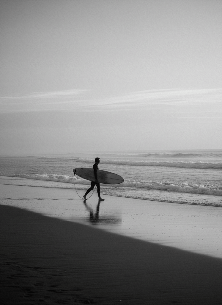 Elegant black and white surf photograph, a surfer walking on the beach at dawn carrying a board, textured wet sand and soft breaking waves behind, minimalist fine-art composition, quiet mood
